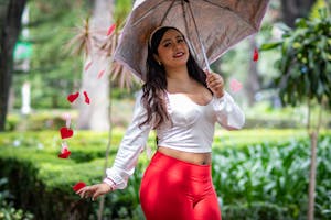 Joyful woman in vibrant attire holding an umbrella adorned with red hearts in a lush outdoor garden.